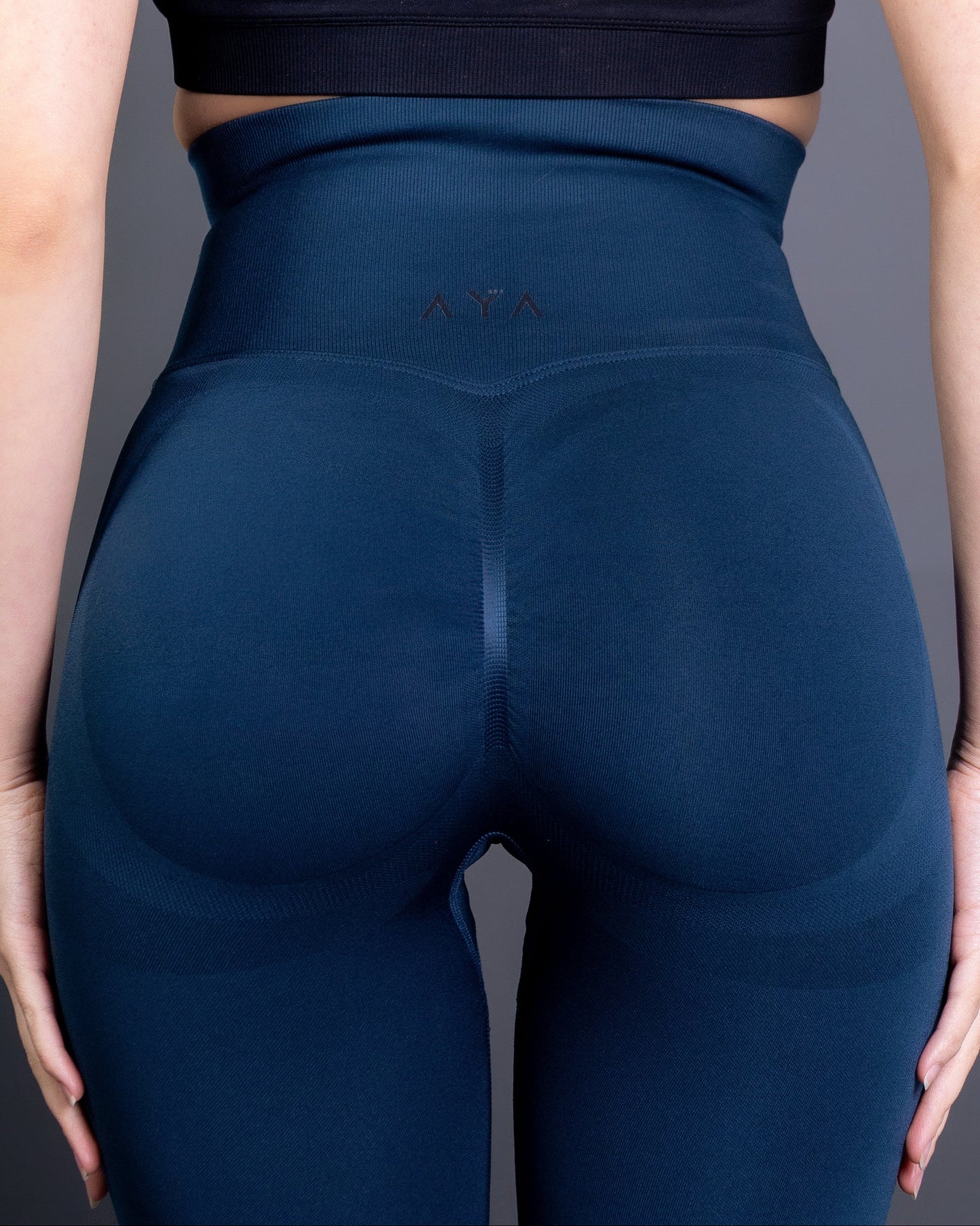 Woman wearing high-waisted, seamless navy blue active leggings paired with a black sports bra and white sneakers. Studio shot against a gray background, showing a confident front-facing pose.