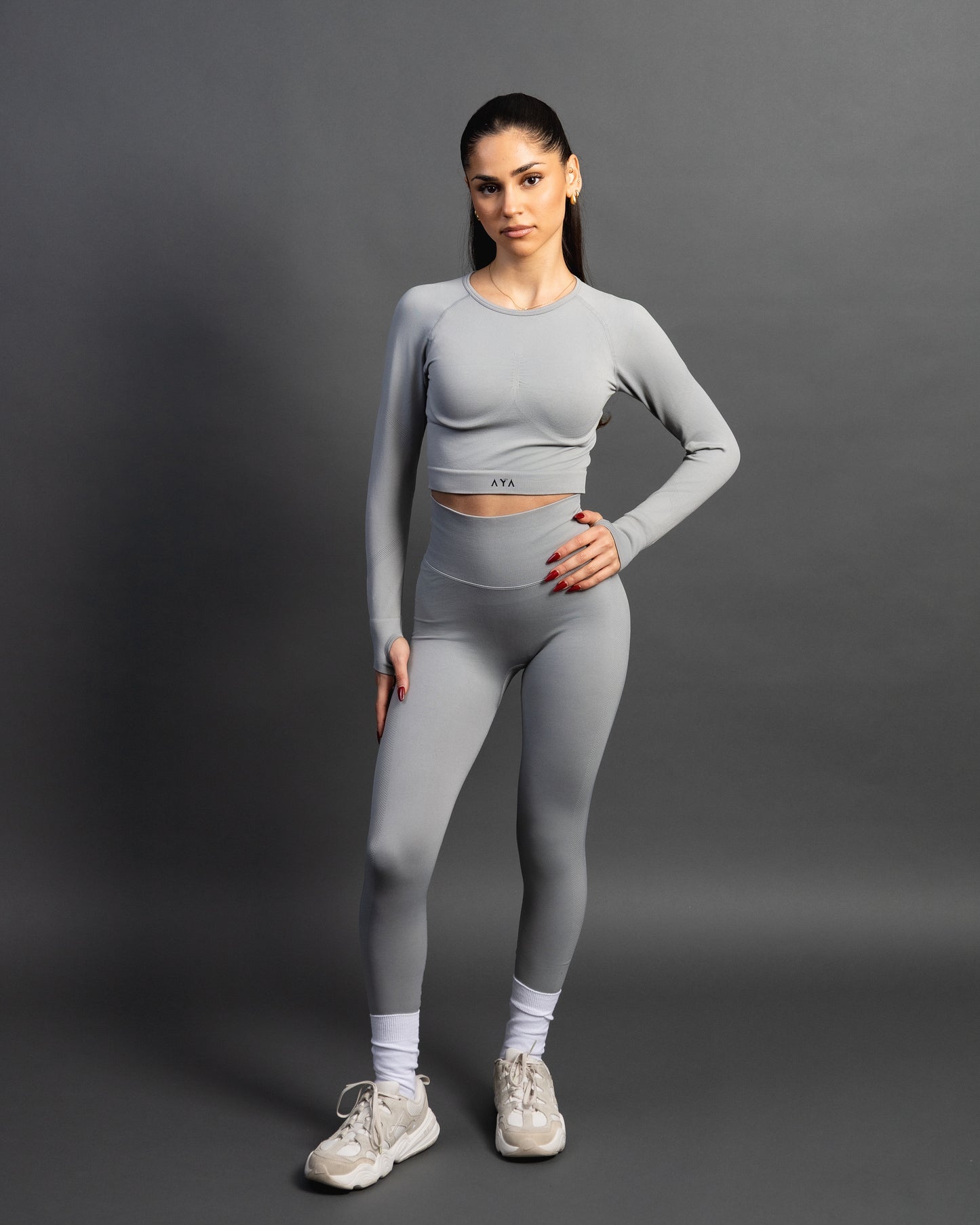 Woman wearing a gray seamless AYA fitness set with long-sleeve crop top and high-waisted leggings, standing in white sneakers in front of a gray studio background.
