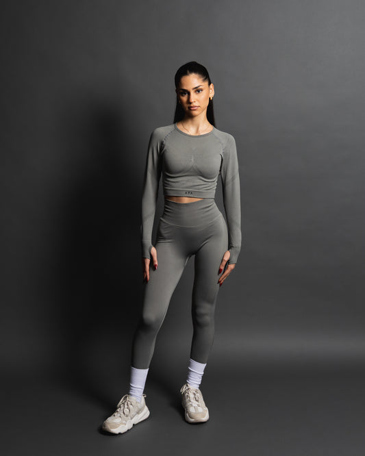 Woman wearing a grey seamless AYA fitness set with long-sleeve crop top and high-waisted leggings, standing in white sneakers in front of a gray studio background.