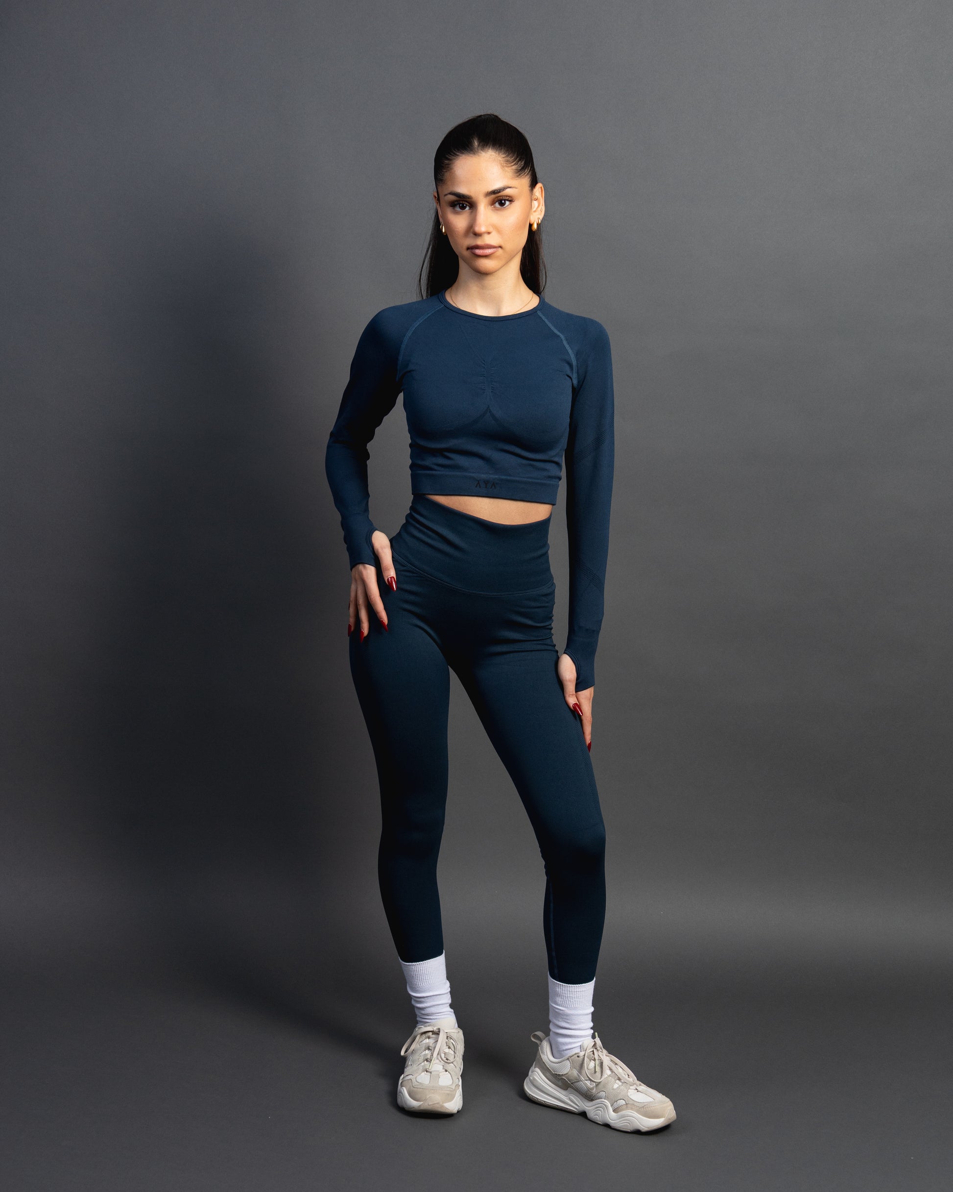 Woman wearing a azurite seamless AYA fitness set with long-sleeve crop top and high-waisted leggings, standing in white sneakers in front of a gray studio background.
