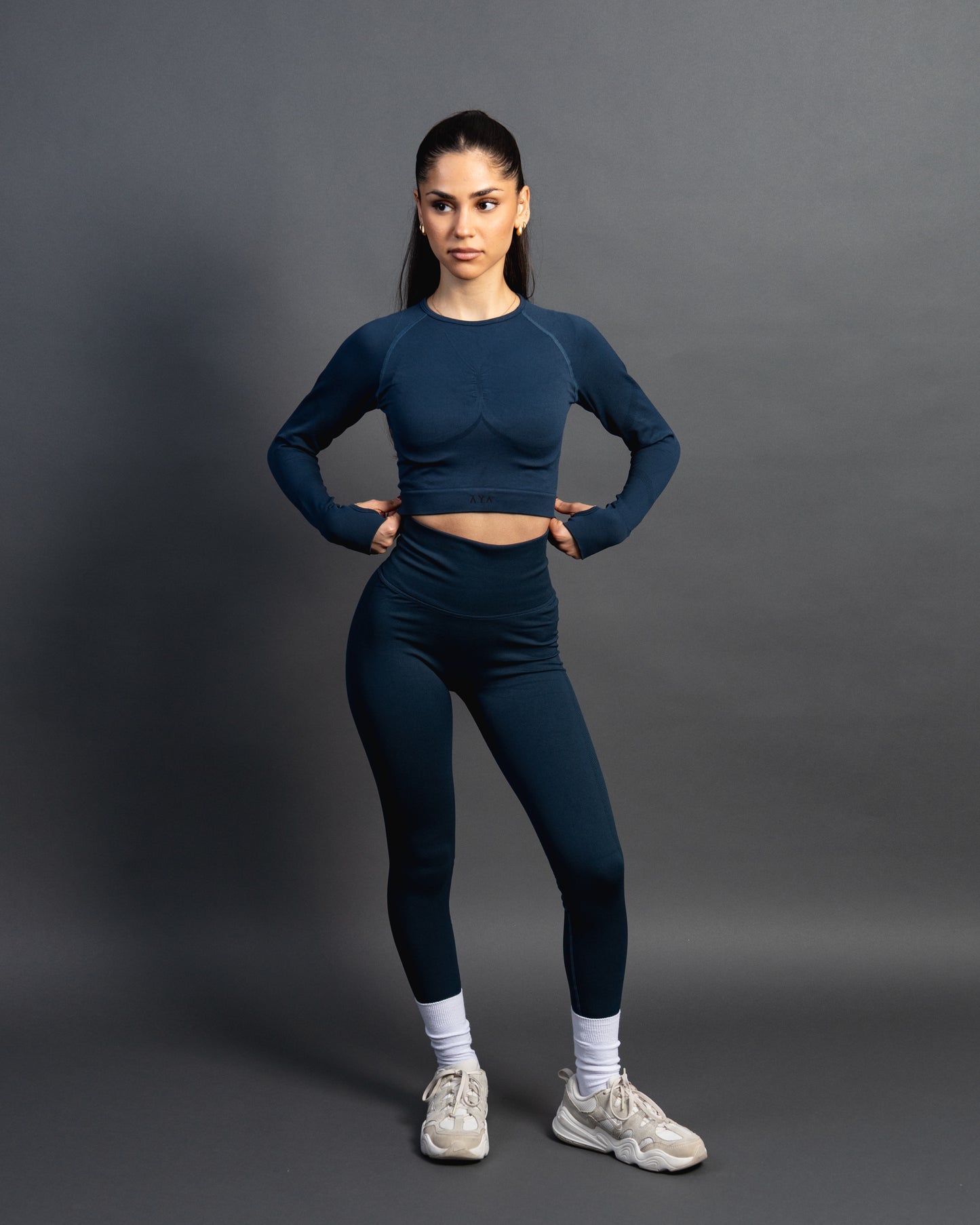 Woman wearing a azurite seamless AYA fitness set with long-sleeve crop top and high-waisted leggings, standing in white sneakers in front of a gray studio background.