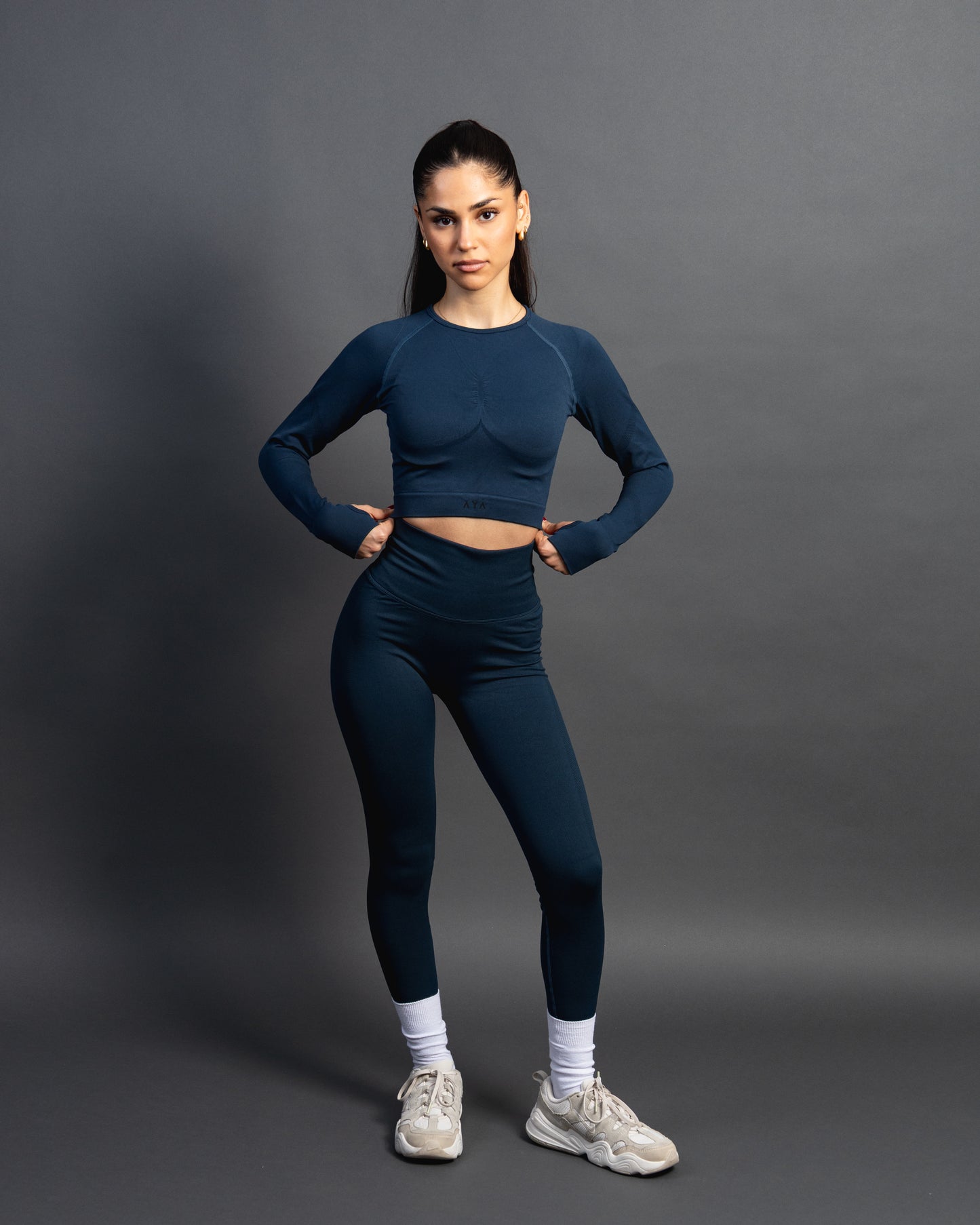 Woman wearing a azurite seamless AYA fitness set with long-sleeve crop top and high-waisted leggings, standing in white sneakers in front of a gray studio background.