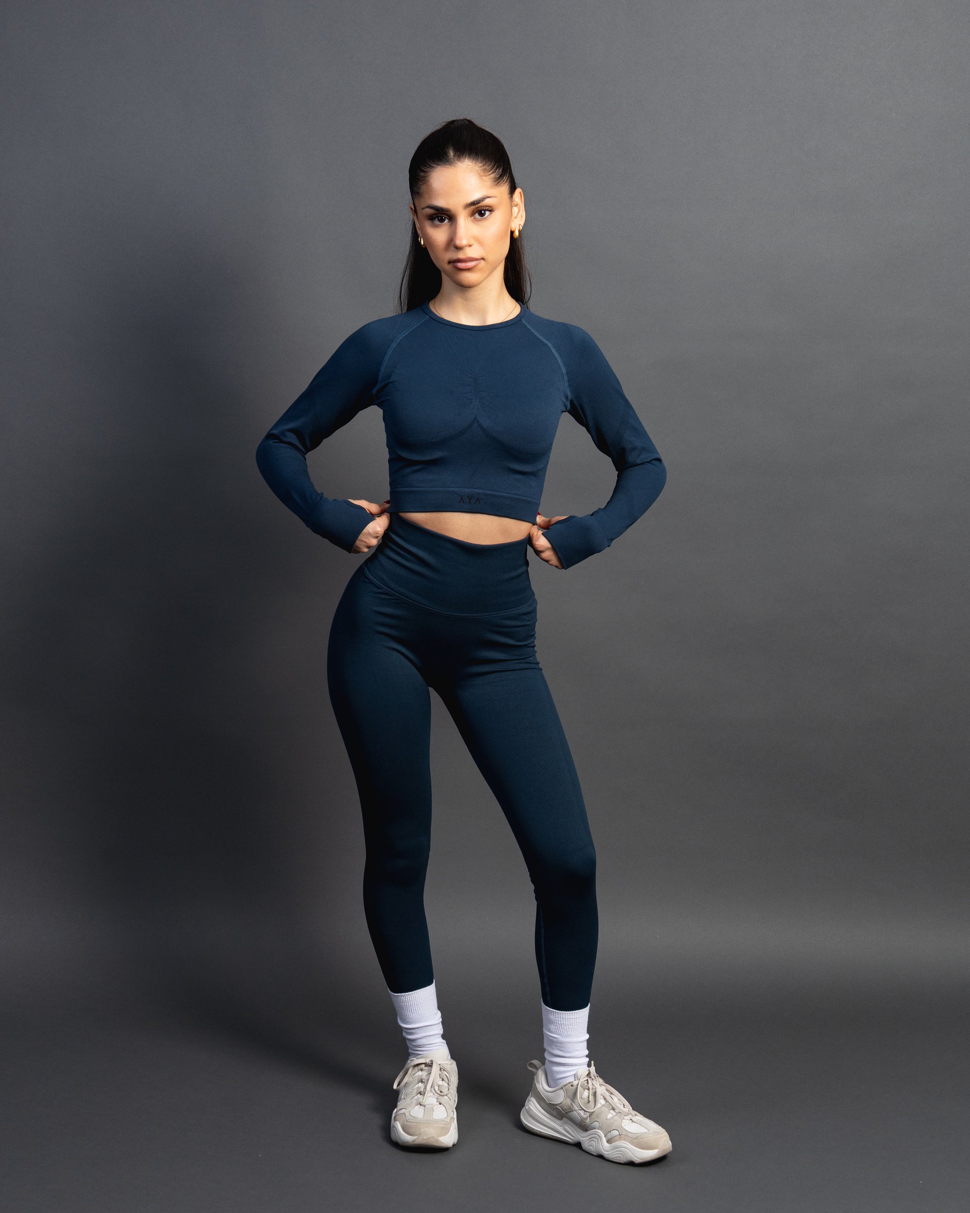 Woman wearing a azurite seamless AYA fitness set with long-sleeve crop top and high-waisted leggings, standing in white sneakers in front of a gray studio background.
