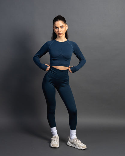 Woman wearing a azurite seamless AYA fitness set with long-sleeve crop top and high-waisted leggings, standing in white sneakers in front of a gray studio background.