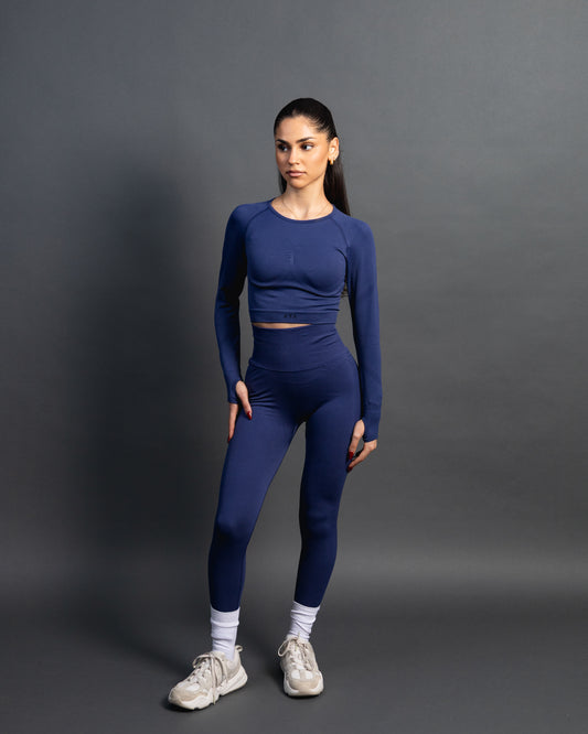 Woman wearing a royal blue seamless AYA fitness set with long-sleeve crop top and high-waisted leggings, standing in white sneakers in front of a gray studio background.
