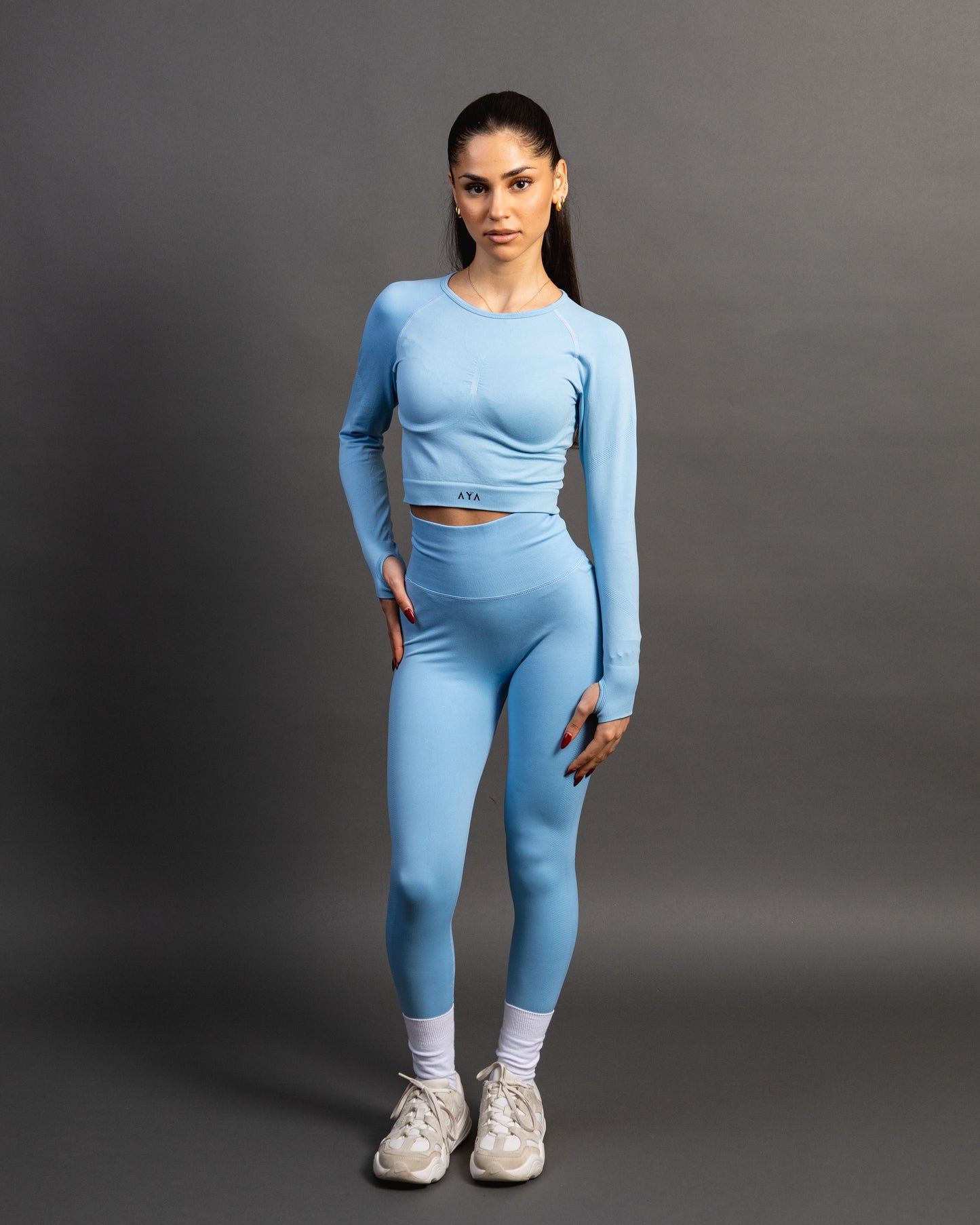 Woman wearing a baby blue seamless AYA fitness set with long-sleeve crop top and high-waisted leggings, standing in white sneakers in front of a gray studio background.