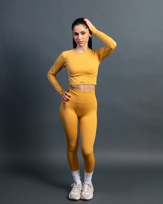 Woman wearing a yellow seamless AYA fitness set with long-sleeve crop top and high-waisted leggings, standing in white sneakers in front of a gray studio background.
