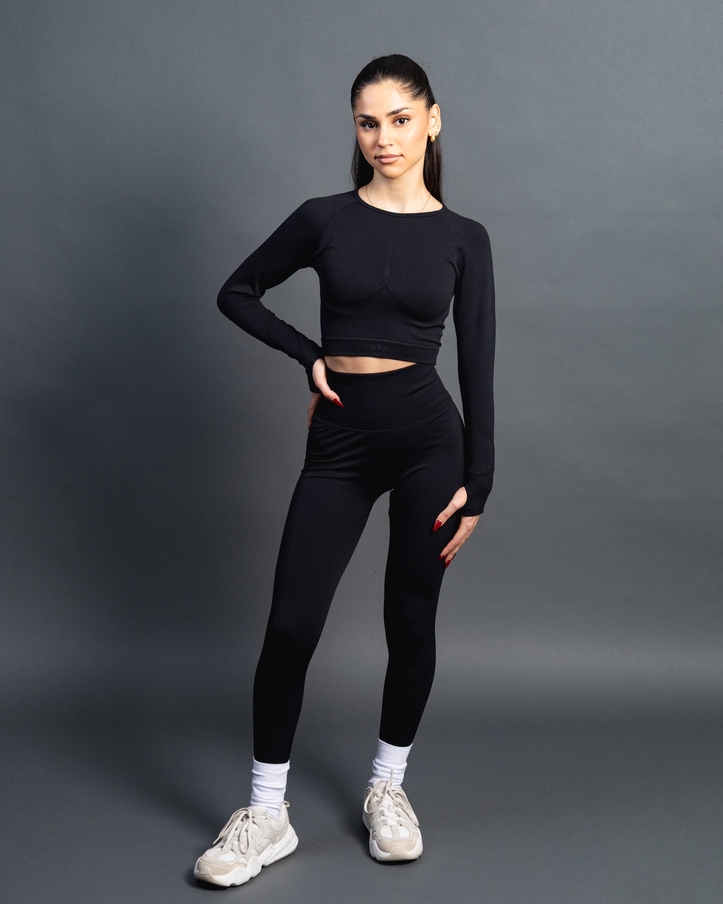 Woman wearing a black seamless AYA fitness set with long-sleeve crop top and high-waisted leggings, standing in white sneakers in front of a gray studio background.