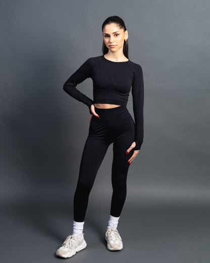 Woman wearing a black seamless AYA fitness set with long-sleeve crop top and high-waisted leggings, standing in white sneakers in front of a gray studio background.