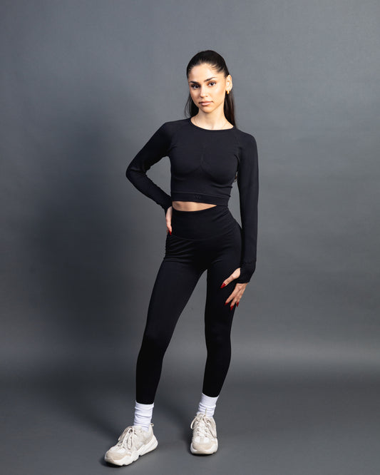 Woman wearing a black seamless AYA fitness set with long-sleeve crop top and high-waisted leggings, standing in white sneakers in front of a gray studio background.