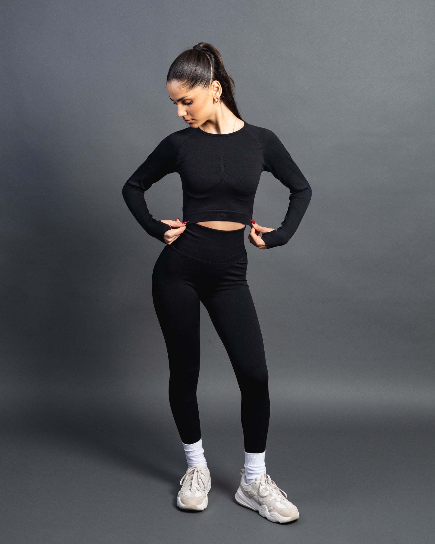 Woman wearing a black seamless AYA fitness set with long-sleeve crop top and high-waisted leggings, standing in white sneakers in front of a gray studio background.