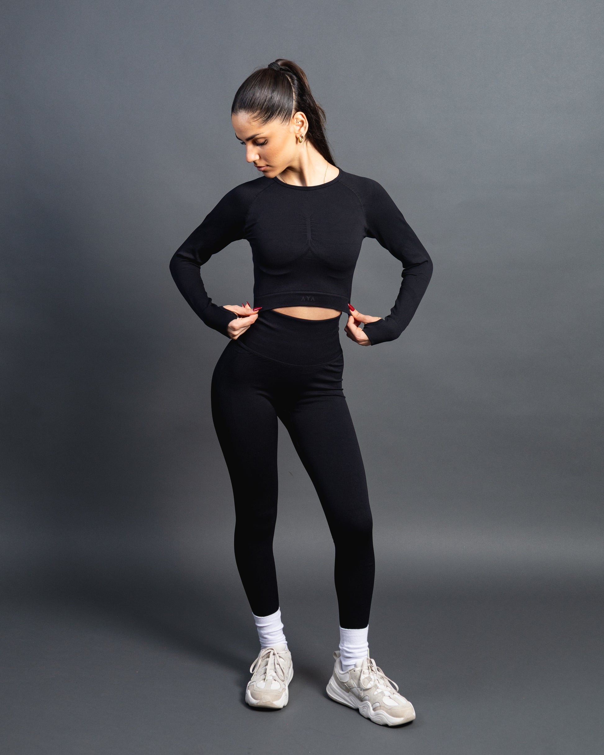 Woman wearing a black seamless AYA fitness set with long-sleeve crop top and high-waisted leggings, standing in white sneakers in front of a gray studio background.