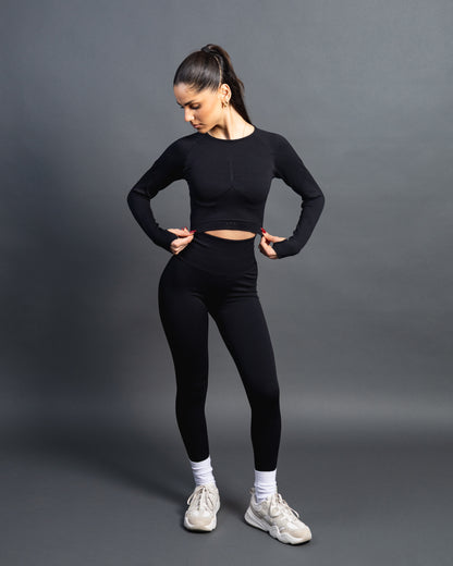Woman wearing a black seamless AYA fitness set with long-sleeve crop top and high-waisted leggings, standing in white sneakers in front of a gray studio background.