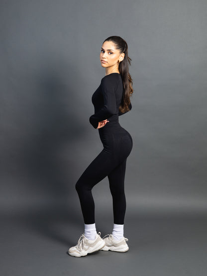 Woman wearing a black seamless AYA fitness set with long-sleeve crop top and high-waisted leggings, standing in white sneakers in front of a gray studio background.
