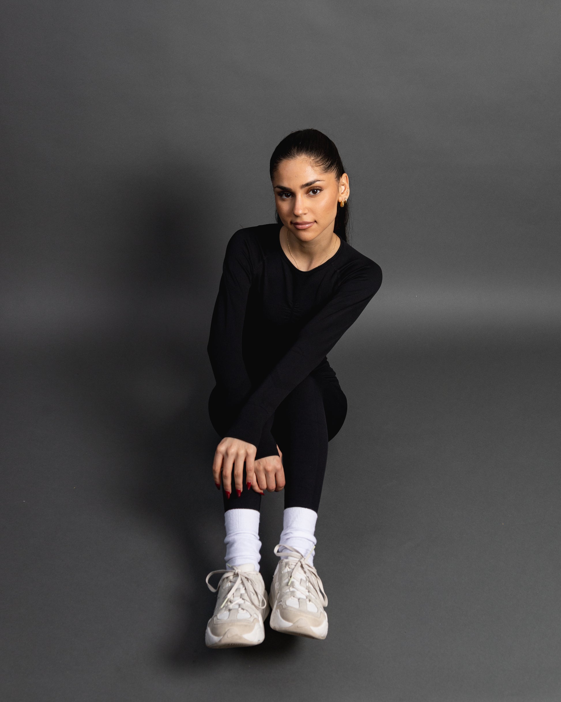 Woman wearing a black seamless AYA fitness set with long-sleeve crop top and high-waisted leggings, standing in white sneakers in front of a gray studio background.