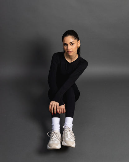 Woman wearing a black seamless AYA fitness set with long-sleeve crop top and high-waisted leggings, standing in white sneakers in front of a gray studio background.
