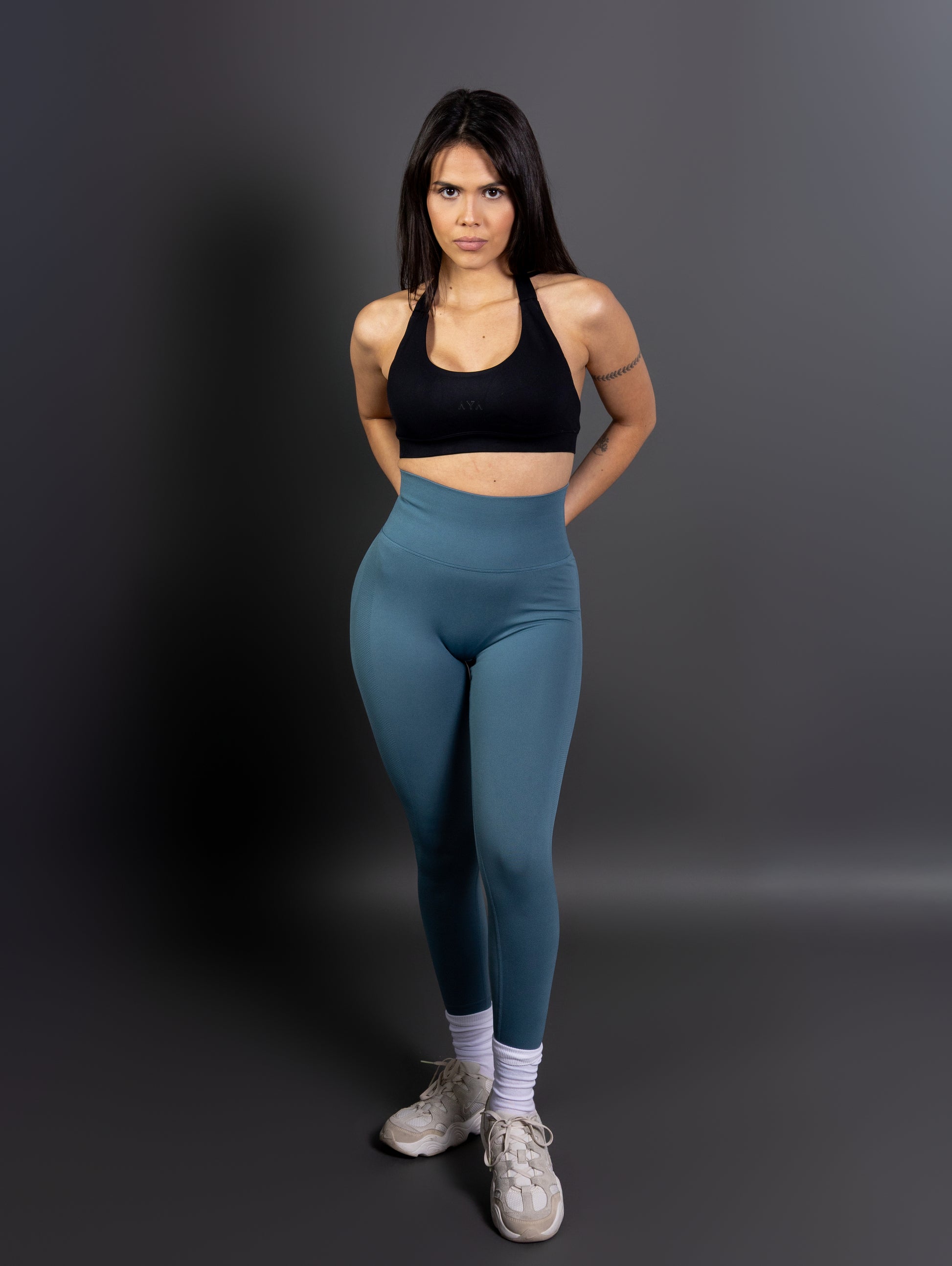 Woman wearing high-waisted, seamless green active leggings paired with a black sports bra and white sneakers. Studio shot against a gray background.