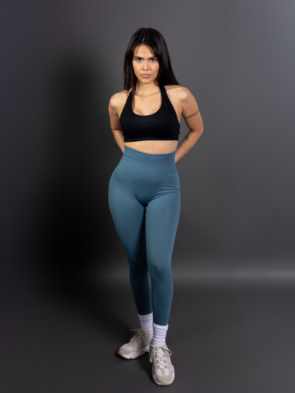 Woman wearing high-waisted, seamless green active leggings paired with a black sports bra and white sneakers. Studio shot against a gray background.