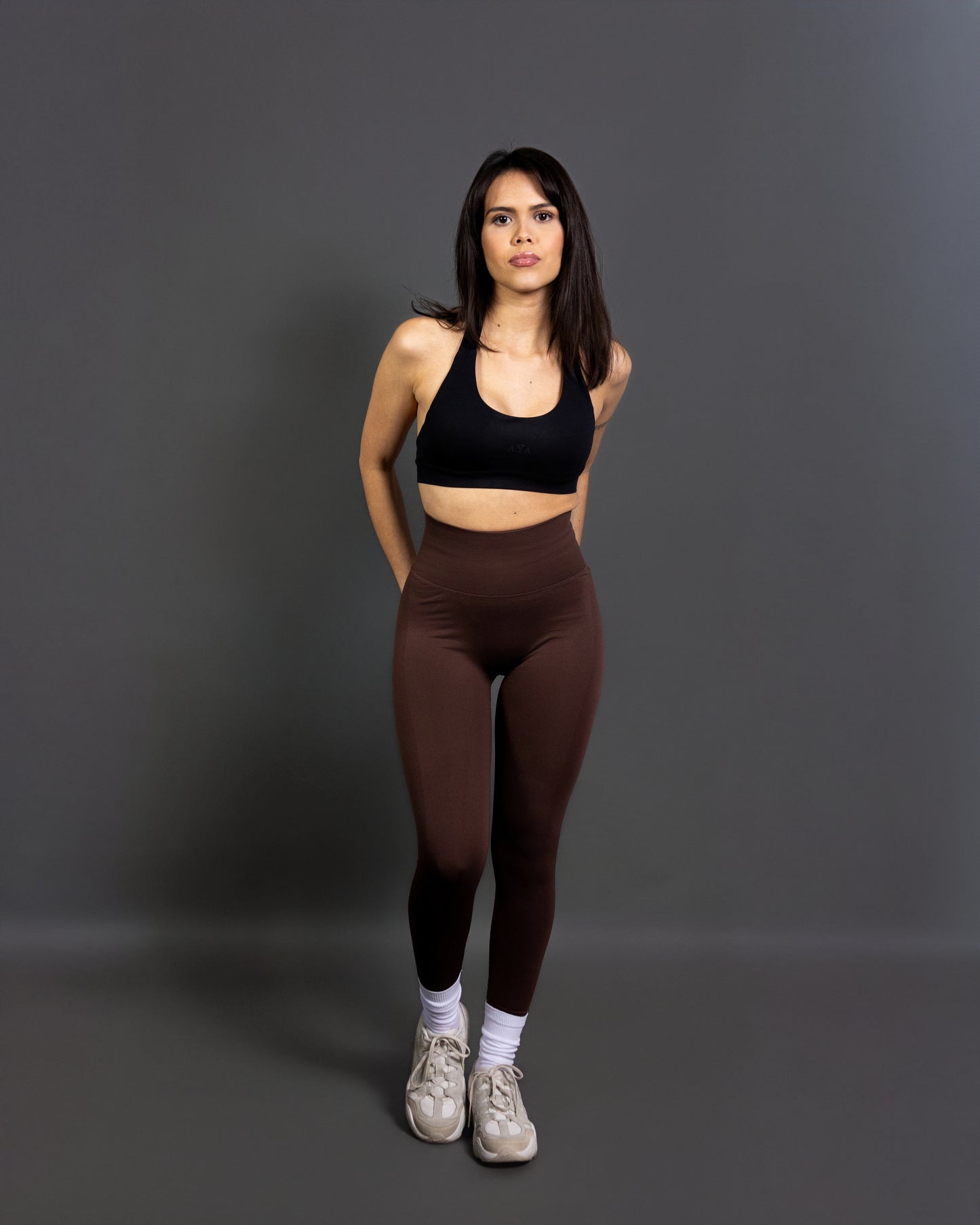 Woman wearing high-waisted, form-fitting brown active leggings with a black patch detail on the left knee, paired with a black sports bra and white sneakers. Studio shot against a gray background.