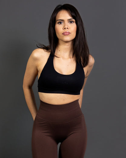 Woman wearing black AYA sports bra and dark brown seamless leggings, standing confidently in a studio setting – modern activewear look with minimalistic design and flattering fit
