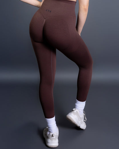 Woman wearing high-waisted, form-fitting brown active leggings with a black patch detail on the left knee, paired with a black sports bra and white sneakers. Studio shot against a gray background.