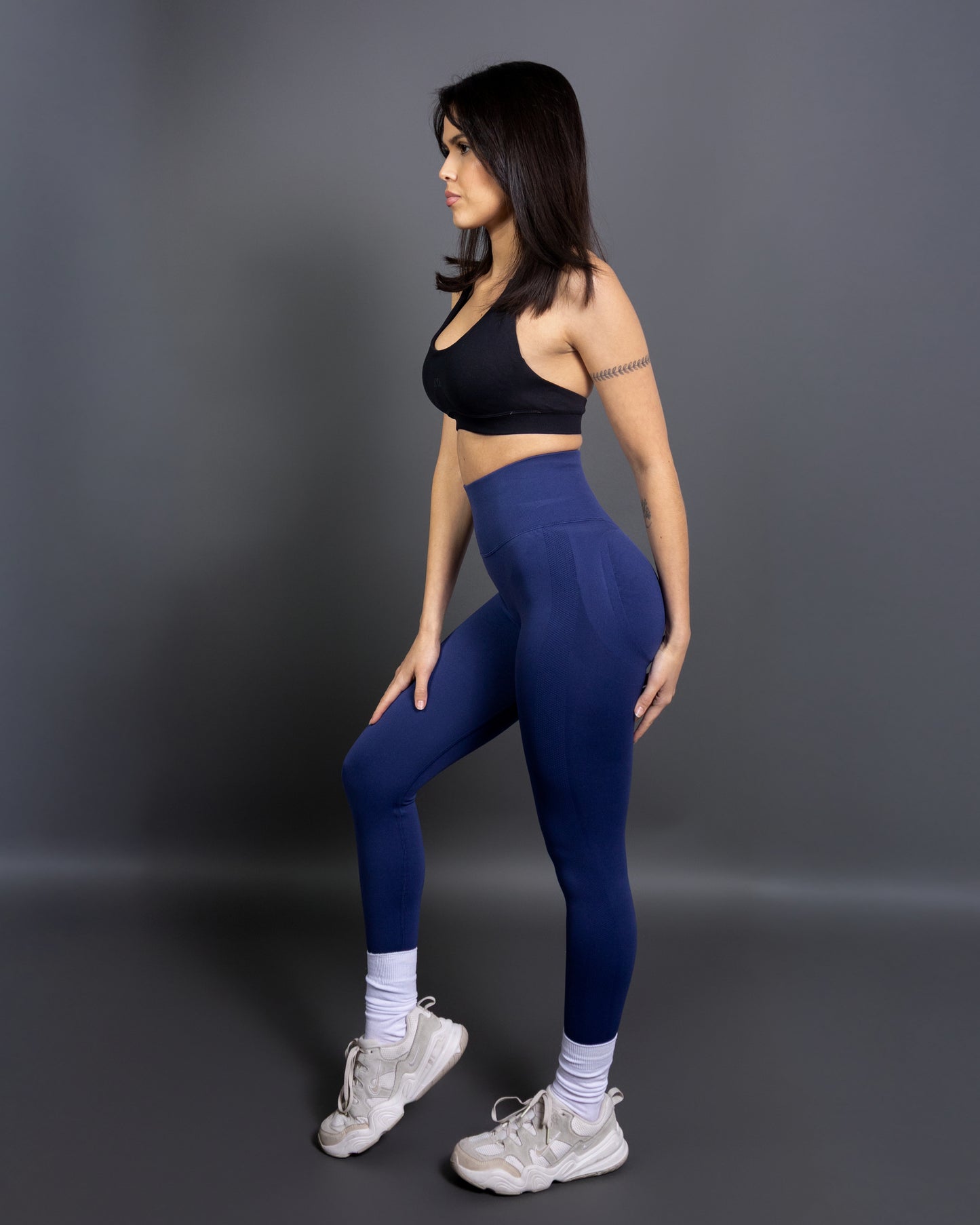 Woman wearing high-waisted, seamless royal blue active leggings paired with a black sports bra and white sneakers. Side profile studio shot against a gray background.