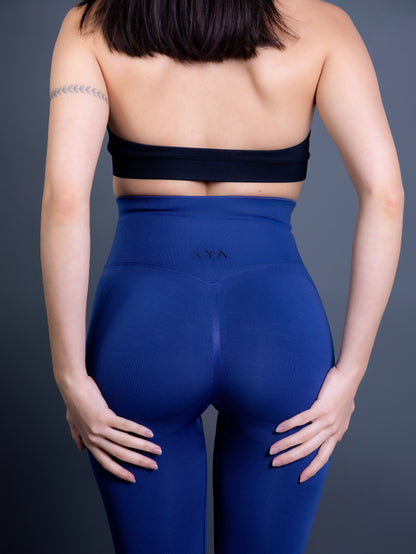 Woman wearing high-waisted, seamless royal blue active leggings paired with a black sports bra and white sneakers. Side profile studio shot against a gray background.