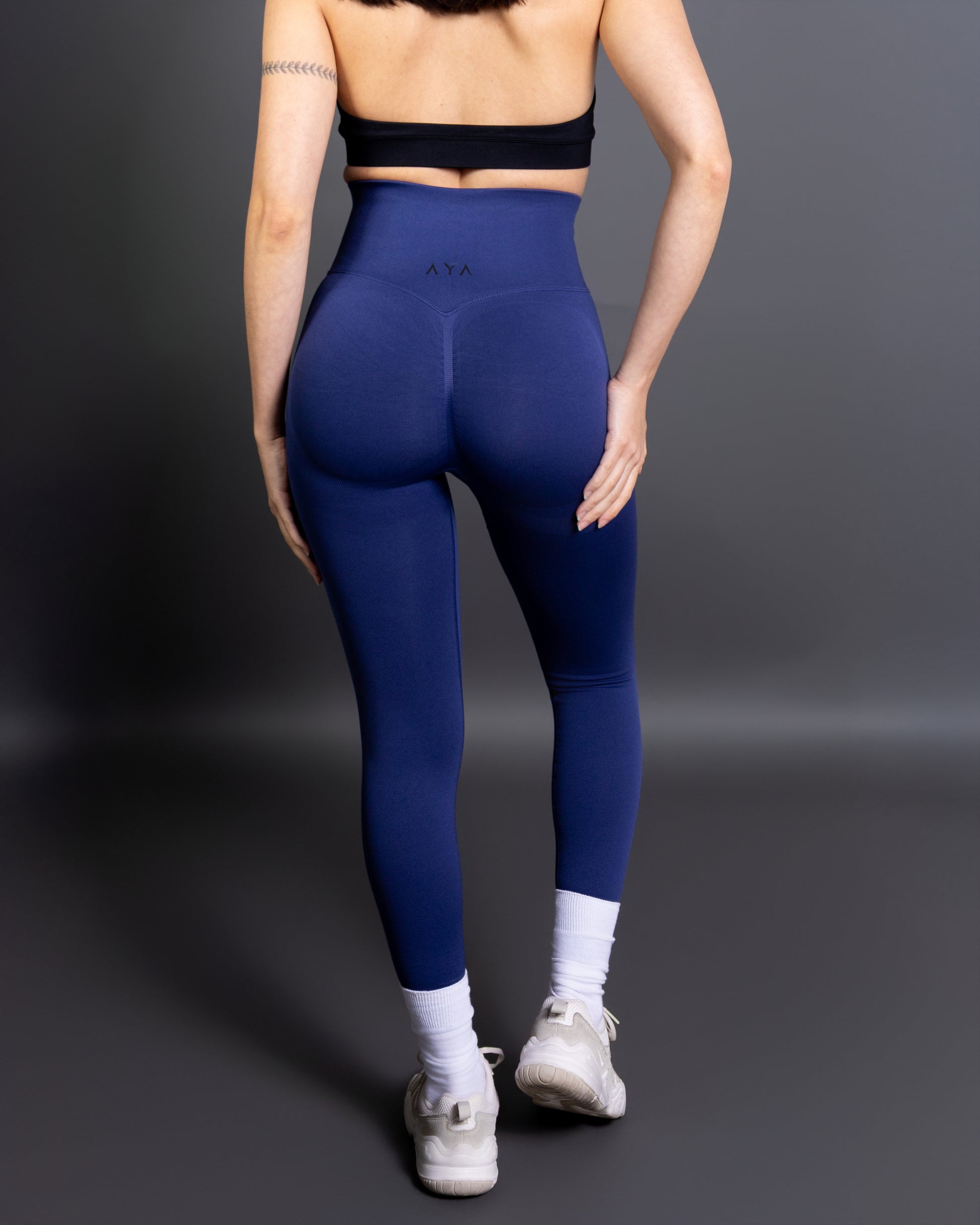 Woman wearing high-waisted, seamless royal blue active leggings paired with a black sports bra and white sneakers. Side profile studio shot against a gray background.
