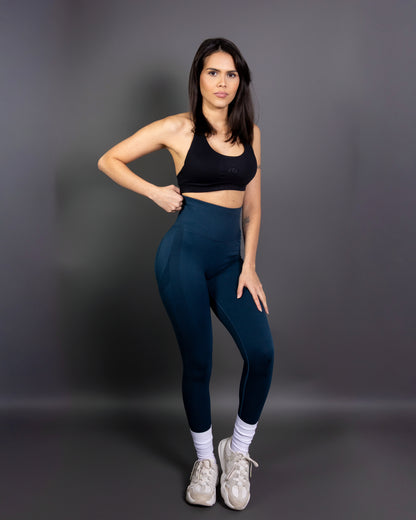 Woman wearing high-waisted, seamless navy blue active leggings paired with a black sports bra and white sneakers. Studio shot against a gray background, showing a confident front-facing pose.