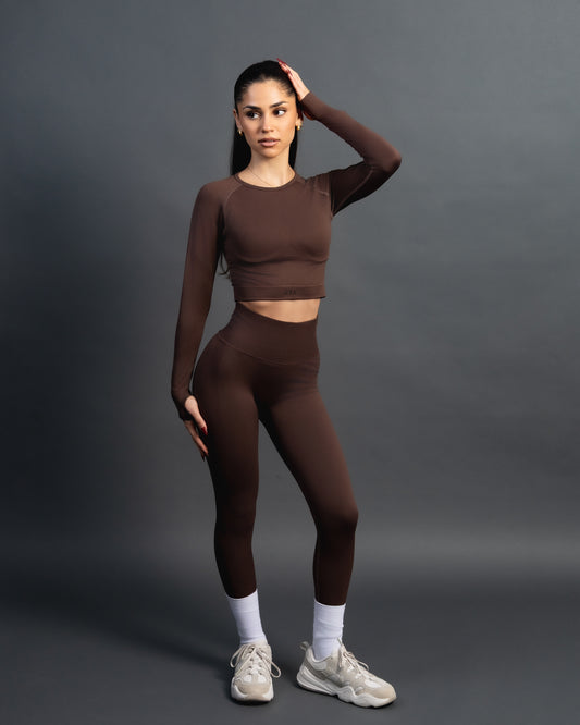Woman wearing a brown seamless AYA fitness set with long-sleeve crop top and high-waisted leggings, standing in white sneakers in front of a gray studio background.