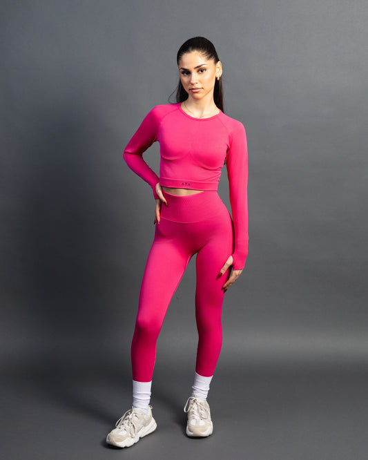 Woman wearing a pink seamless AYA fitness set with long-sleeve crop top and high-waisted leggings, standing in white sneakers in front of a gray studio background.