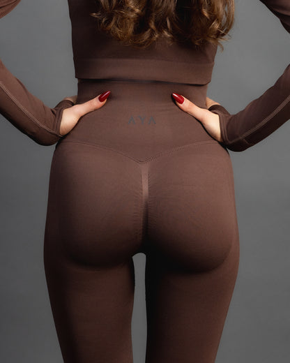 Woman wearing high-waisted, form-fitting brown active leggings with a black patch detail on the left knee, paired with a black sports bra and white sneakers. Studio shot against a gray background.