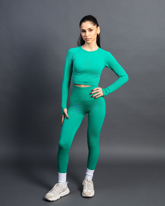 Woman wearing a green seamless AYA fitness set with long-sleeve crop top and high-waisted leggings, standing in white sneakers in front of a gray studio background.