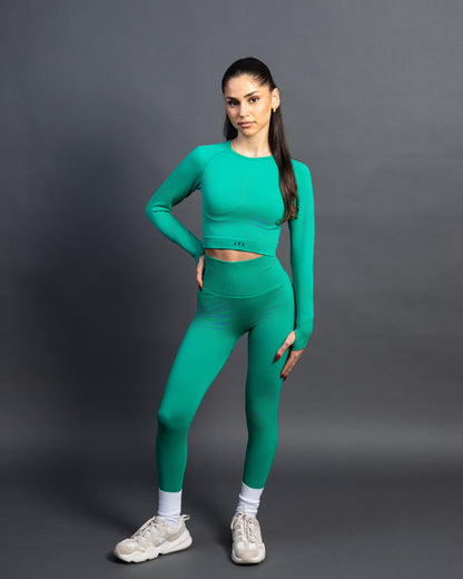 Woman wearing a green seamless AYA fitness set with long-sleeve crop top and high-waisted leggings, standing in white sneakers in front of a gray studio background.