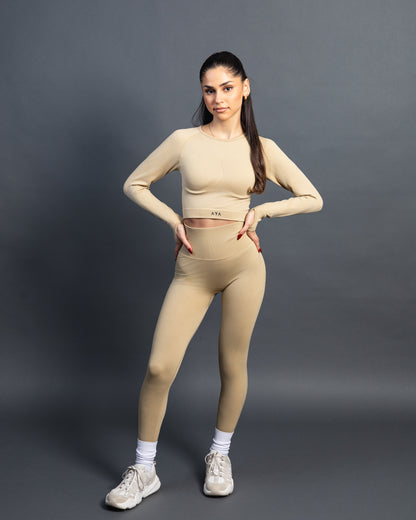 Woman wearing a beige seamless AYA fitness set with long-sleeve crop top and high-waisted leggings, standing in white sneakers in front of a gray studio background.