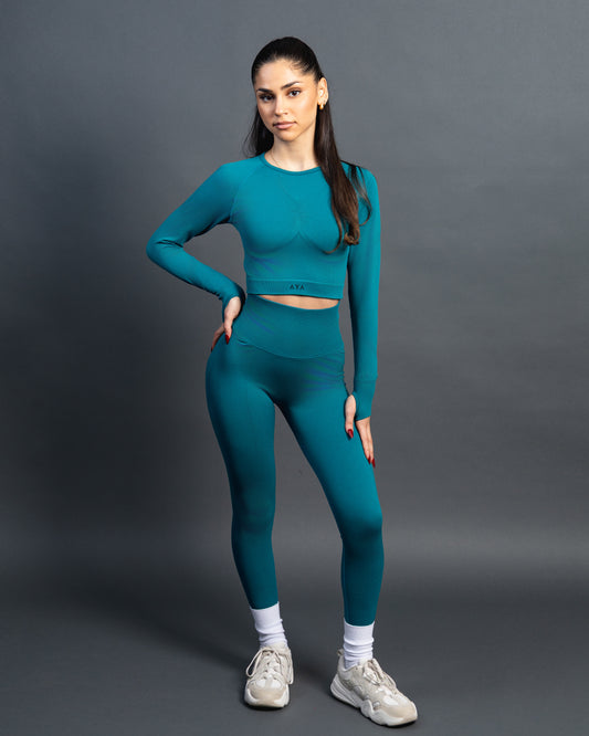 Woman wearing a green seamless AYA fitness set with long-sleeve crop top and high-waisted leggings, standing in white sneakers in front of a gray studio background.