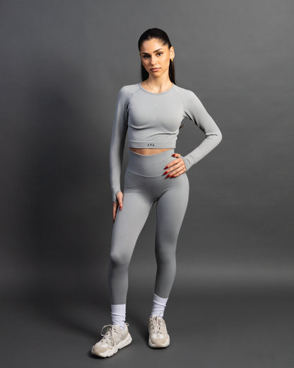 Woman wearing a gray seamless AYA fitness set with long-sleeve crop top and high-waisted leggings, standing in white sneakers in front of a gray studio background.