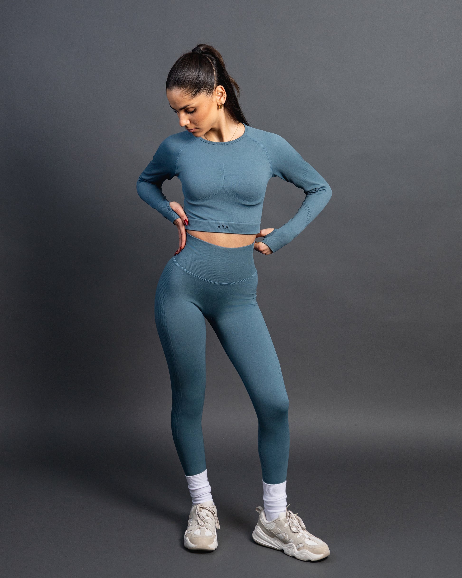 Woman wearing a green seamless AYA fitness set with long-sleeve crop top and high-waisted leggings, standing in white sneakers in front of a gray studio background.