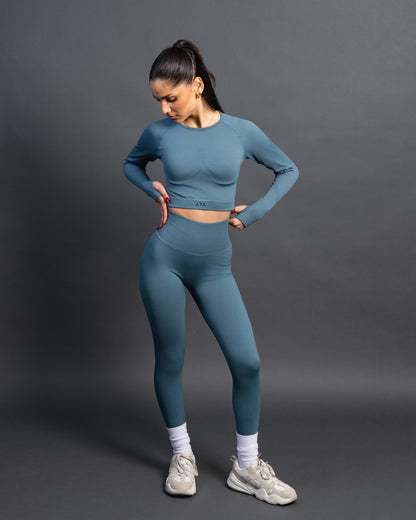 Woman wearing a green seamless AYA fitness set with long-sleeve crop top and high-waisted leggings, standing in white sneakers in front of a gray studio background.