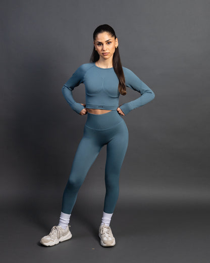 Woman wearing a green seamless AYA fitness set with long-sleeve crop top and high-waisted leggings, standing in white sneakers in front of a gray studio background.