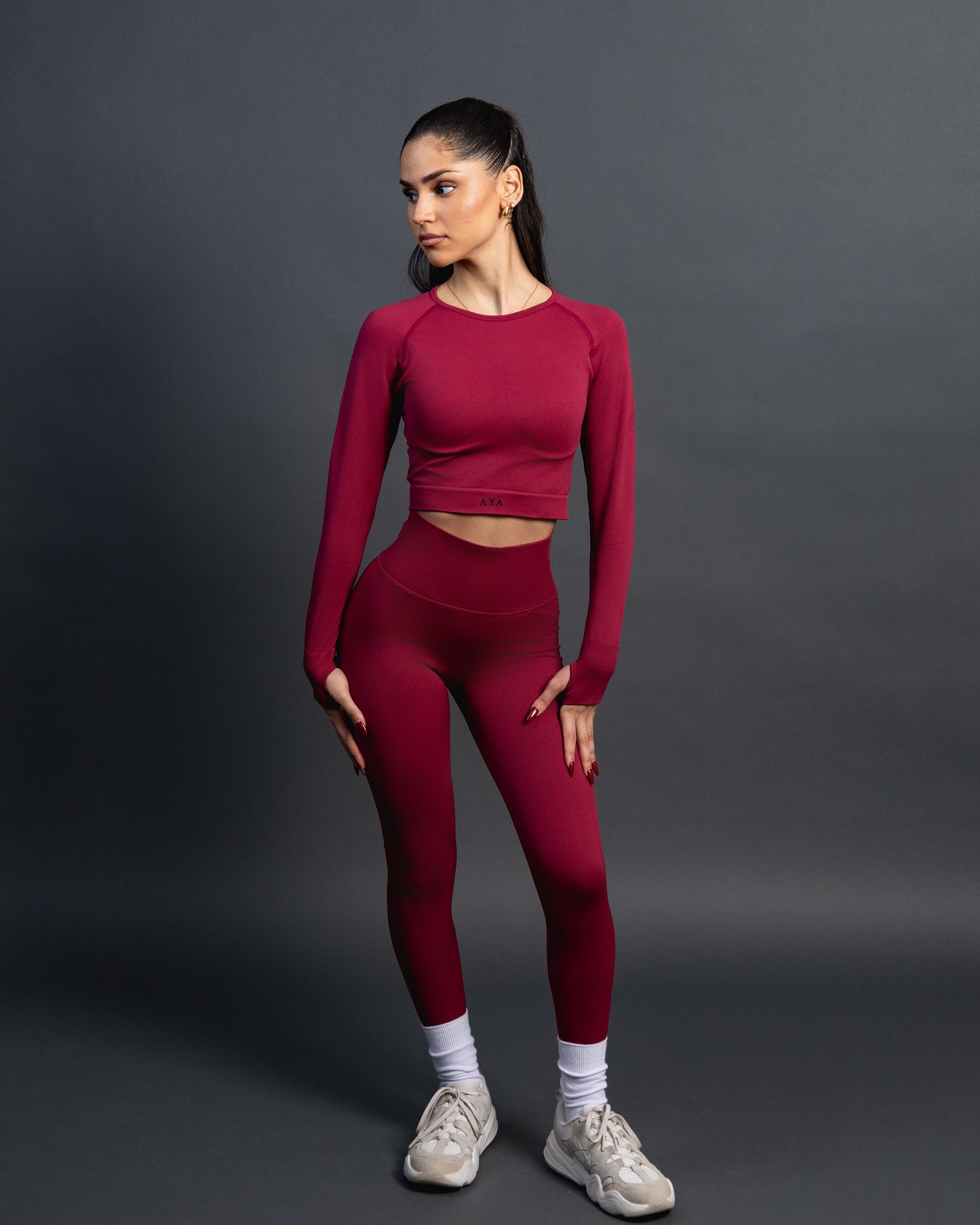 Woman wearing a red seamless AYA fitness set with long-sleeve crop top and high-waisted leggings, standing in white sneakers in front of a gray studio background.