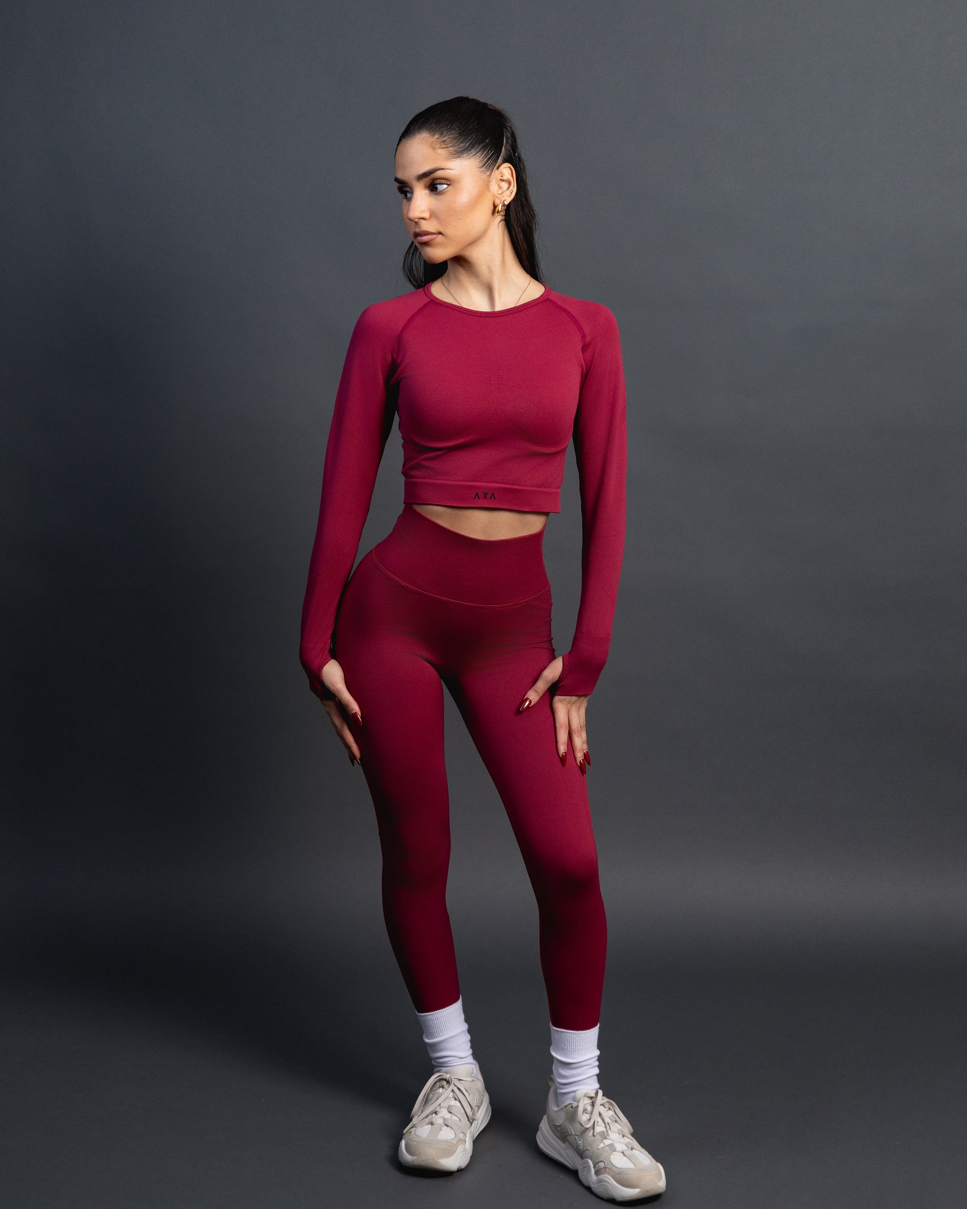 Woman wearing a red seamless AYA fitness set with long-sleeve crop top and high-waisted leggings, standing in white sneakers in front of a gray studio background.