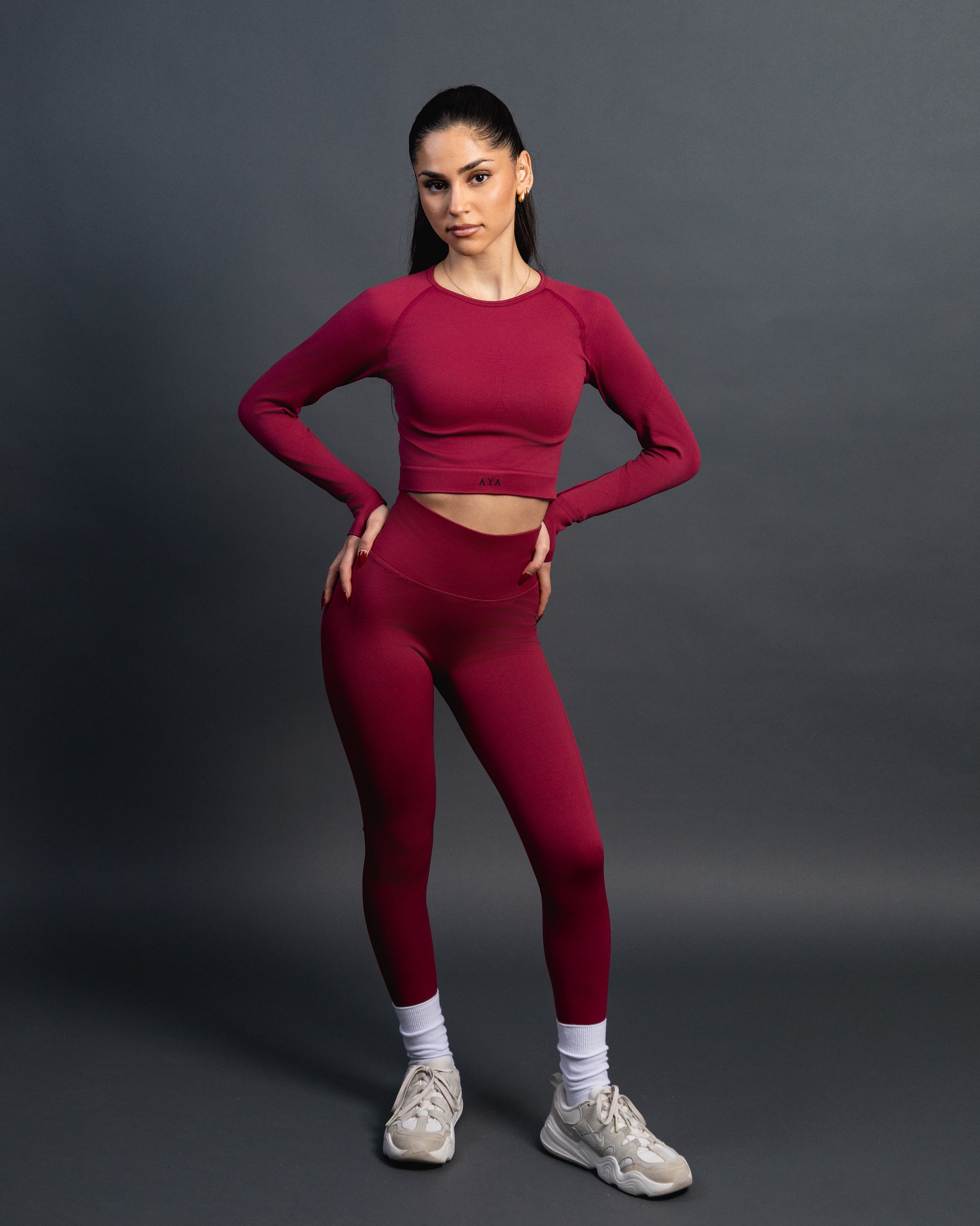 Woman wearing a red seamless AYA fitness set with long-sleeve crop top and high-waisted leggings, standing in white sneakers in front of a gray studio background.