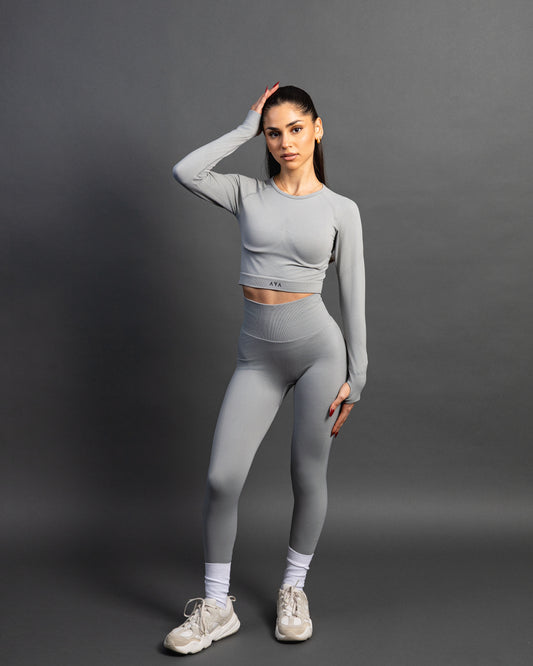 Woman wearing a gray seamless AYA fitness set with long-sleeve crop top and high-waisted leggings, standing in white sneakers in front of a gray studio background.