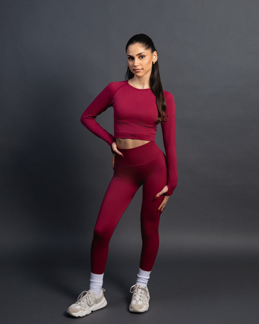 Woman wearing a red seamless AYA fitness set with long-sleeve crop top and high-waisted leggings, standing in white sneakers in front of a gray studio background.