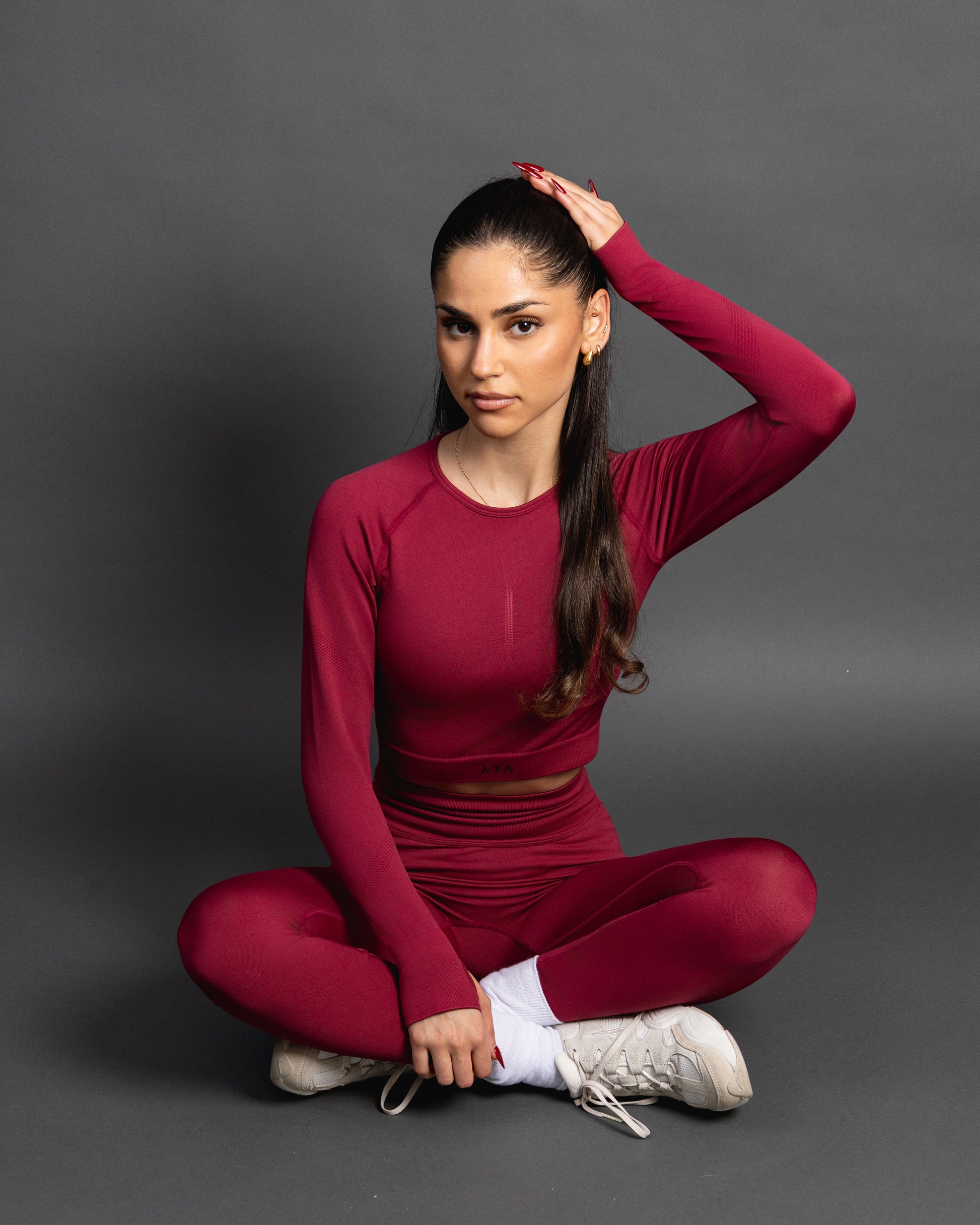Woman wearing a red seamless AYA fitness set with long-sleeve crop top and high-waisted leggings, standing in white sneakers in front of a gray studio background.