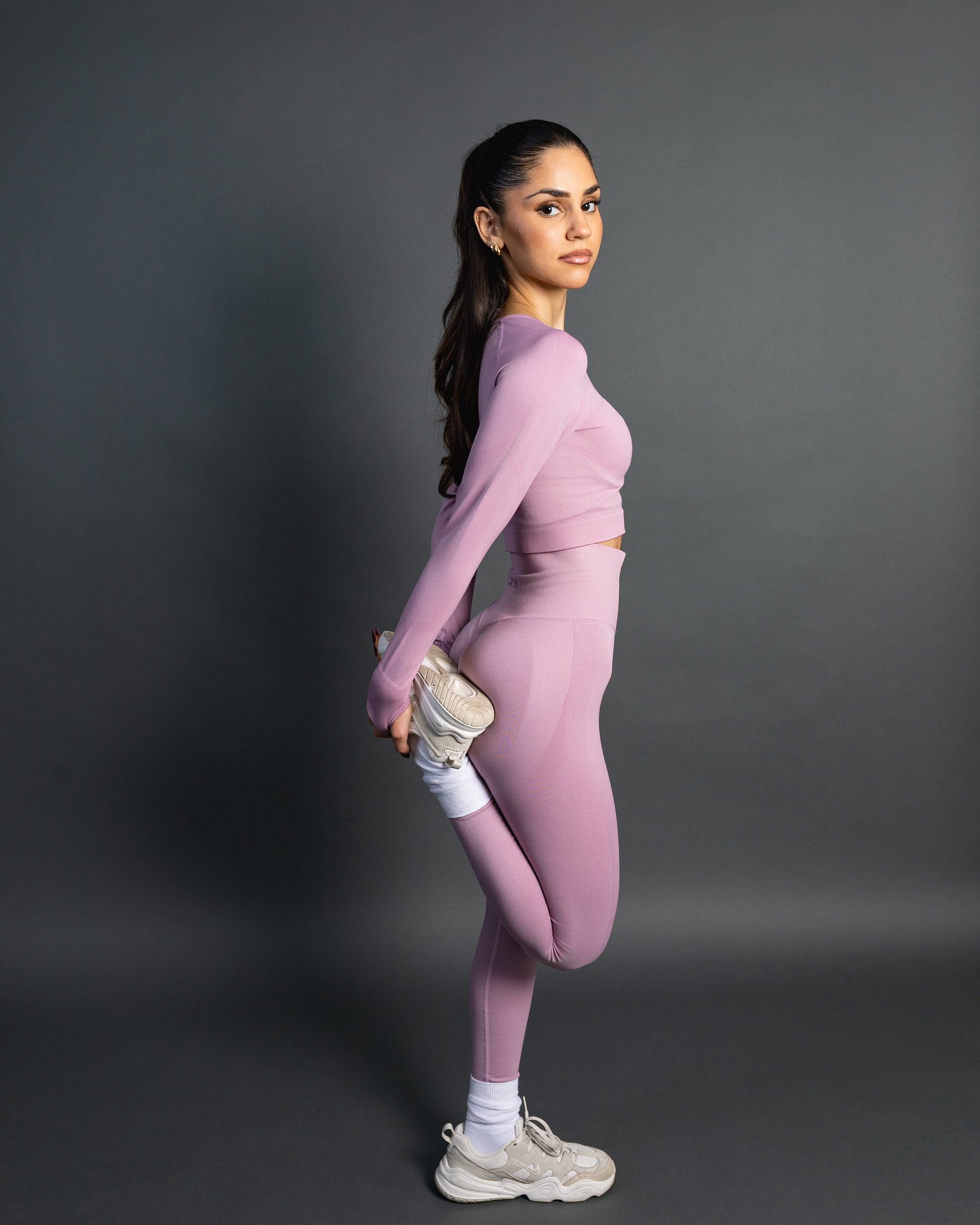 Woman wearing a baby pink seamless AYA fitness set with long-sleeve crop top and high-waisted leggings, standing in white sneakers in front of a gray studio background.