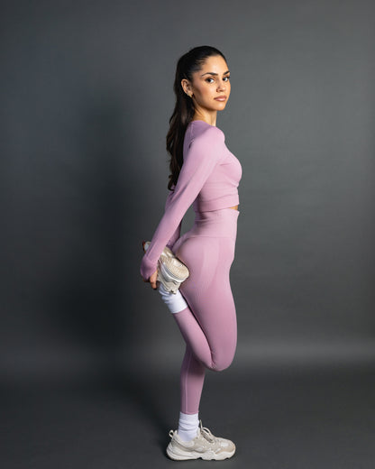 Woman wearing a baby pink seamless AYA fitness set with long-sleeve crop top and high-waisted leggings, standing in white sneakers in front of a gray studio background.