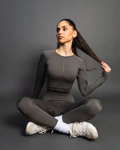Woman wearing a gray seamless AYA fitness set with long-sleeve crop top and high-waisted leggings, standing in white sneakers in front of a gray studio background.